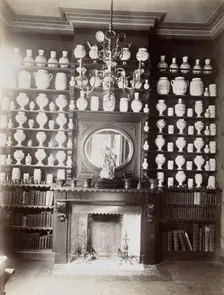 Drug jars and books on shelves surrounding a mantlepiece with a statue and a mirror, c1900.. Creator: Unknown