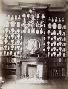Drug jars and books on shelves surrounding a mantlepiece with a statue and a mirror, c1900.. Creator: Unknown