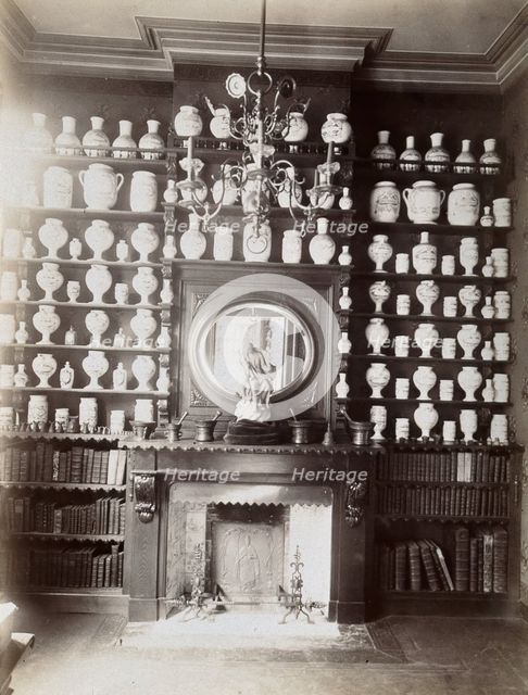 Drug jars and books on shelves surrounding a mantlepiece with a statue and a mirror, c1900.. Creator: Unknown.