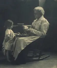 Elderly woman in a rocking chair peeling apple with young girl standing in front of her, c1900. Creator: Elizabeth B. Brownell