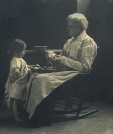 Elderly woman in a rocking chair peeling apple with young girl standing in front of her, c1900. Creator: Elizabeth B. Brownell