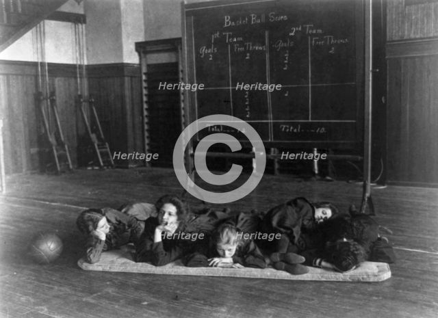 Five girls lying on mat in gymnasium in front of basketball scoreboard..., Washington, D.C., (1899?) Creator: Frances Benjamin Johnston.