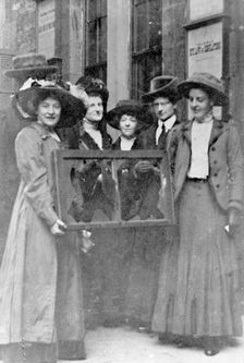 Five suffragettes holding a broken window in its frame, 1912