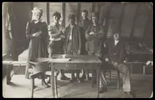 French prisoners of war, one in drag, gather round a small wooden table, [between 1910 and 1919?]. Creator: Unknown