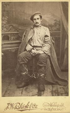 Full-length portrait of a man in convict clothing and leg irons, seated..., between 1880 and 1886. Creator: Unknown