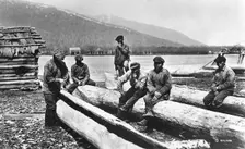 Kamchadals on the river bank near the baht and logs prepared for hollowing out the baht, 1922-1923. Creator: Rene Malaise
