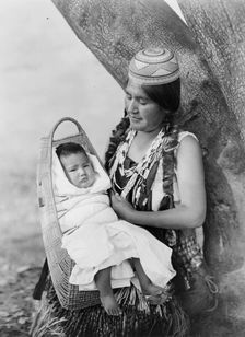 Hupa mother, three-quarter length portrait, seated, facing left, holding baby, c1923. Creator: Edward Sheriff Curtis