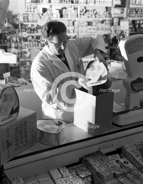 Shopkeeper unpacking canned gammon joints, Mexborough, South Yorkshire, 1963.  Creator: Michael Walters.