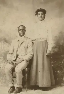 Studio portrait of a young couple, he seated, she with hand on his shoulder, c1900. Creator: Carbon Studio