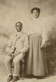 Studio portrait of a young couple, he seated, she with hand on his shoulder, c1900. Creator: Carbon Studio