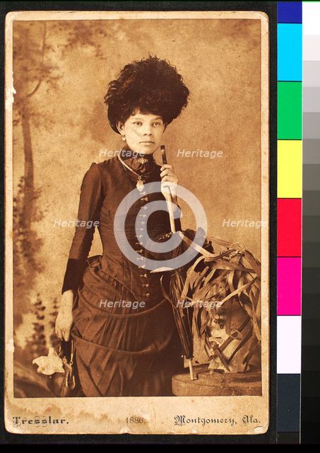 Standing woman, wearing dress with bustle, holding purse at side and umbrella resting..., 1886. Creator: Unknown.