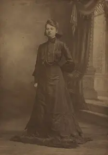 Portrait of a young woman in long black dress, standing, about 1900. Creator: George W Killian