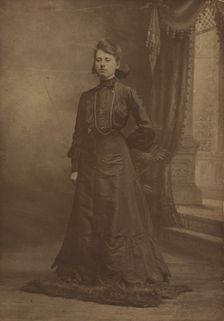 Portrait of a young woman in long black dress, standing, about 1900. Creator: George W Killian