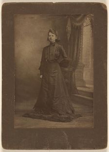 Portrait of a young woman in long black dress, standing, about 1900. Creator: George W Killian