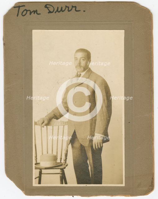 Photograph of Tom Durr standing next to a chair with a hat on it, ca. 1910. Creator: Unknown.