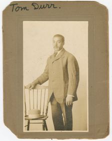 Photograph of Tom Durr standing next to a chair with a hat on it, ca. 1910. Creator: Unknown