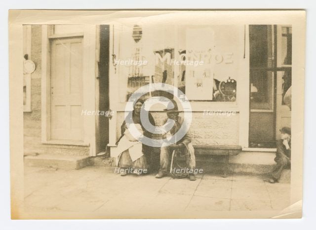 Photograph of a man and woman sitting outside of a storefront, early 20th century. Creator: Unknown.