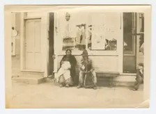 Photograph of a man and woman sitting outside of a storefront, early 20th century. Creator: Unknown