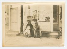 Photograph of a man and woman sitting outside of a storefront, early 20th century. Creator: Unknown