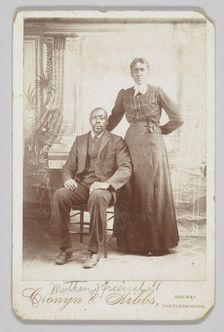 Photograph of a man sitting down with a woman standing next to him, 1880s - 1900. Creator: Cronyn & Hibbs