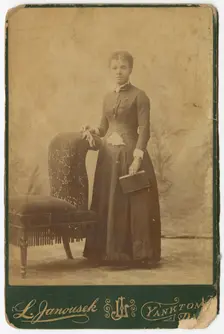 Photograph of a woman standing next to a chair and holding a book, late 19th century. Creator: Louis Janousek