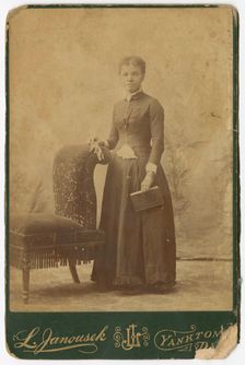Photograph of a woman standing next to a chair and holding a book, late 19th century. Creator: Louis Janousek