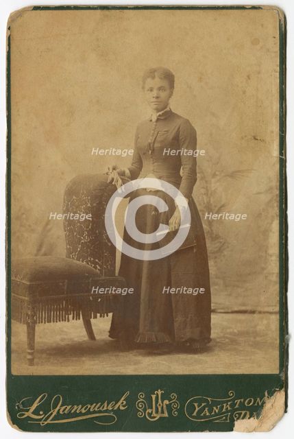 Photograph of a woman standing next to a chair and holding a book, late 19th century. Creator: Louis Janousek.