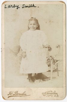 Photograph of Larby Smith standing next to a wicker chair., late 19th century. Creator: Henry Bruhn