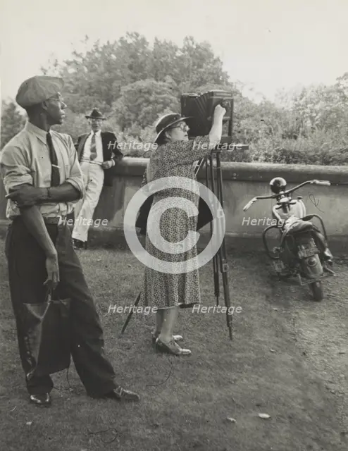 Photographer Frances Benjamin Johnston standing beside her view camera preparing to take..., 1938. Creator: Unknown.