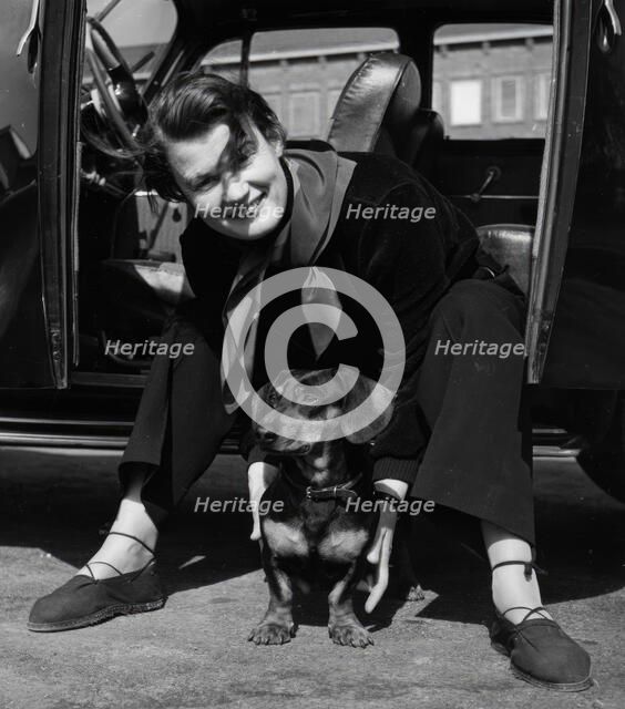 Photographer Kerstin Bernhard sits in the doorway of her car and holds a dachshund, 1955-1960. Creator: Unknown.