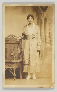 Photographic postcard of a woman standing next to a wooden chair, 1904-1918. Creator: Unknown
