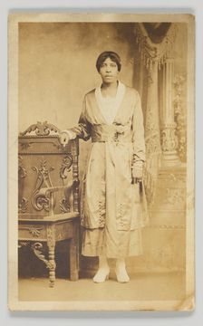 Photographic postcard of a woman standing next to a wooden chair, 1904-1918. Creator: Unknown