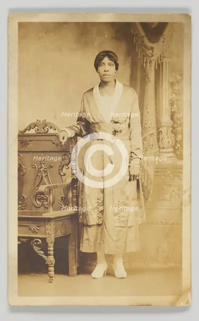 Photographic postcard of a woman standing next to a wooden chair, 1904-1918. Creator: Unknown.