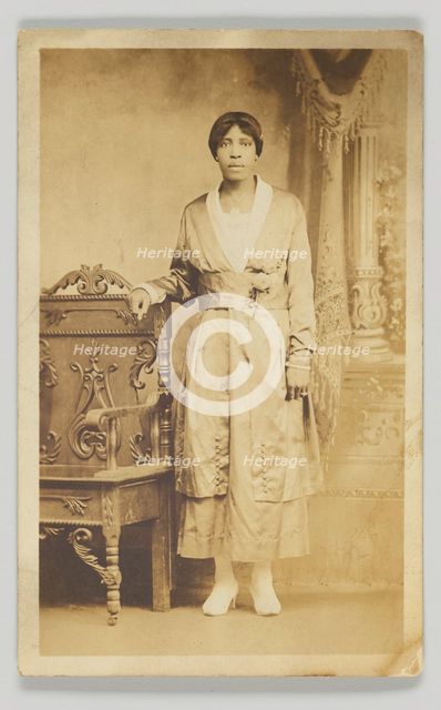 Photographic postcard of a woman standing next to a wooden chair, 1904-1918. Creator: Unknown.