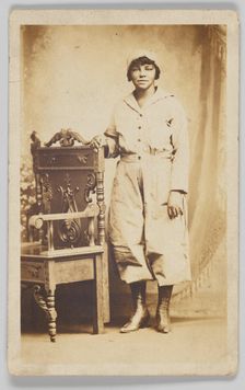 Photographic postcard of a woman standing next to a wooden chair, 1918-1930. Creator: Unknown
