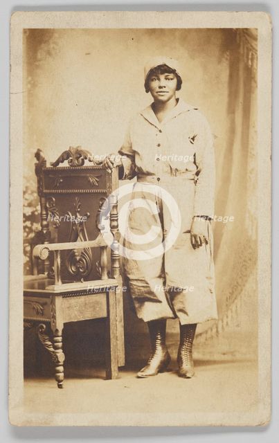 Photographic postcard of a woman standing next to a wooden chair, 1918-1930. Creator: Unknown.