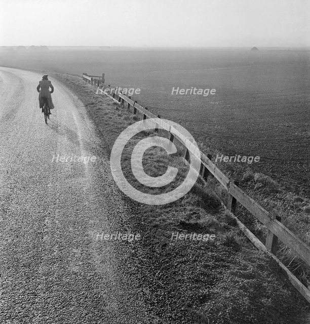 Woman cycling along a road raised up above the surrounding fenland, Cambridgeshire, early 1950s. Artist: John Gay.
