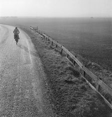 Woman cycling along a road raised up above the surrounding fenland, Cambridgeshire, early 1950s. Artist: John Gay