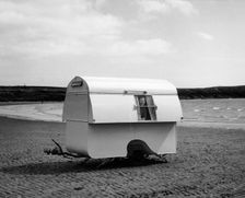 Woman smiling from the window of a trailer caravan on a beach, (c1960s?)