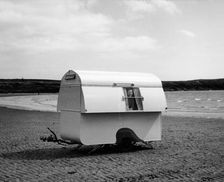 Woman smiling from the window of a trailer caravan on a beach, (c1960s?)