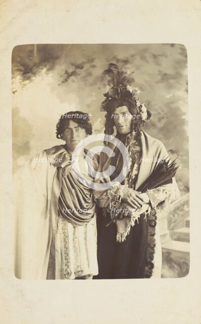 Two men in drag, one wearing a detailed feathered head garment, pose for a portrait, [1900-1909?]. Creator: Frank Wood.