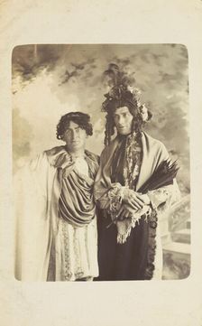 Two men in drag, one wearing a detailed feathered head garment, pose for a portrait, [1900-1909?]. Creator: Frank Wood