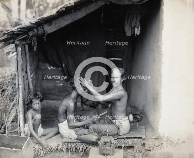 Two men sitting cross-legged; one is a barber shaving the front of the other's head, c1900. Creator: Unknown.