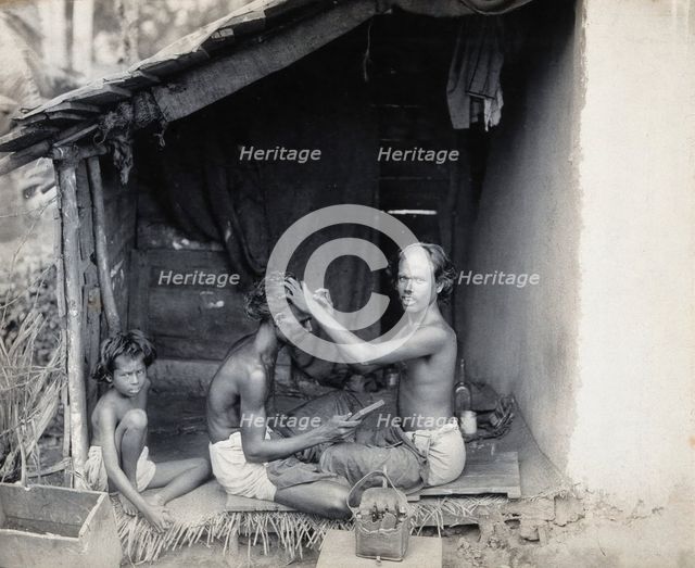 Two men sitting cross-legged; one is a barber shaving the front of the other's head, c1900. Creator: Unknown.