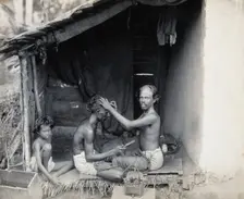 Two men sitting cross-legged; one is a barber shaving the front of the other's head, c1900. Creator: Unknown