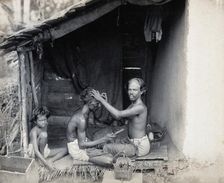 Two men sitting cross-legged; one is a barber shaving the front of the other's head, c1900. Creator: Unknown
