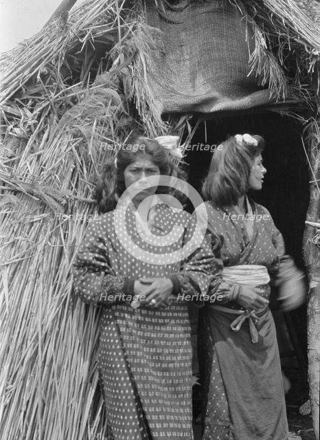 Two Ainu women standing outside the entrance of a hut, 1908. Creator: Arnold Genthe.