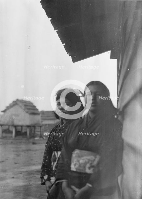 Two Ainu women standing outside by the wall of a wooden hut, 1908. Creator: Arnold Genthe.