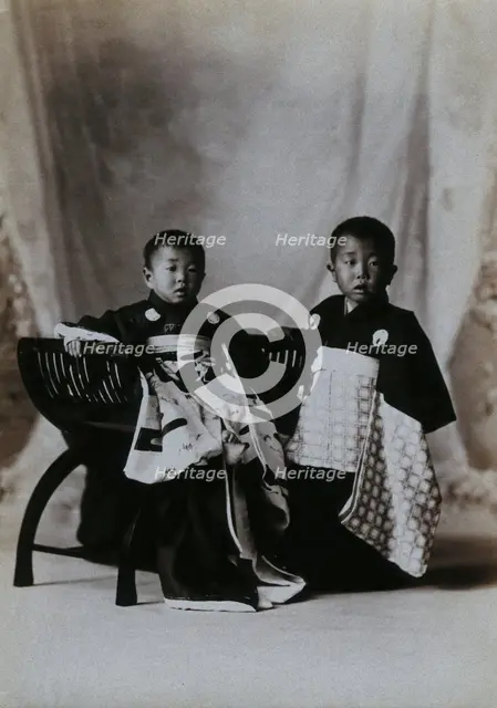 Two boys in a photographic studio, one seated on a chair, in front of a curtained backdrop, c1900. Creator: Unknown.