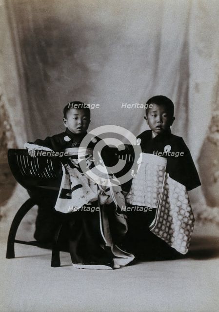 Two boys in a photographic studio, one seated on a chair, in front of a curtained backdrop, c1900. Creator: Unknown.